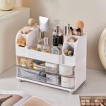 Heaime white acrylic makeup organizer with two drawers and an open top section holding brushes, lipsticks, and skincare bottl