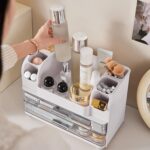 Heaime white acrylic makeup organizer with two drawers and an open top section holding brushes, lipsticks, and skincare bottl